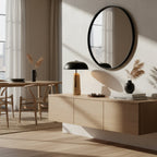A Clérachic table lamp on a floating oak cabinet beneath an oval black mirror, with a dining table and sheer curtains behind.