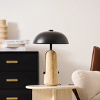 A Clérachic table lamp on a round travertine side table in front of a black chest with brass handles and a cream chair.