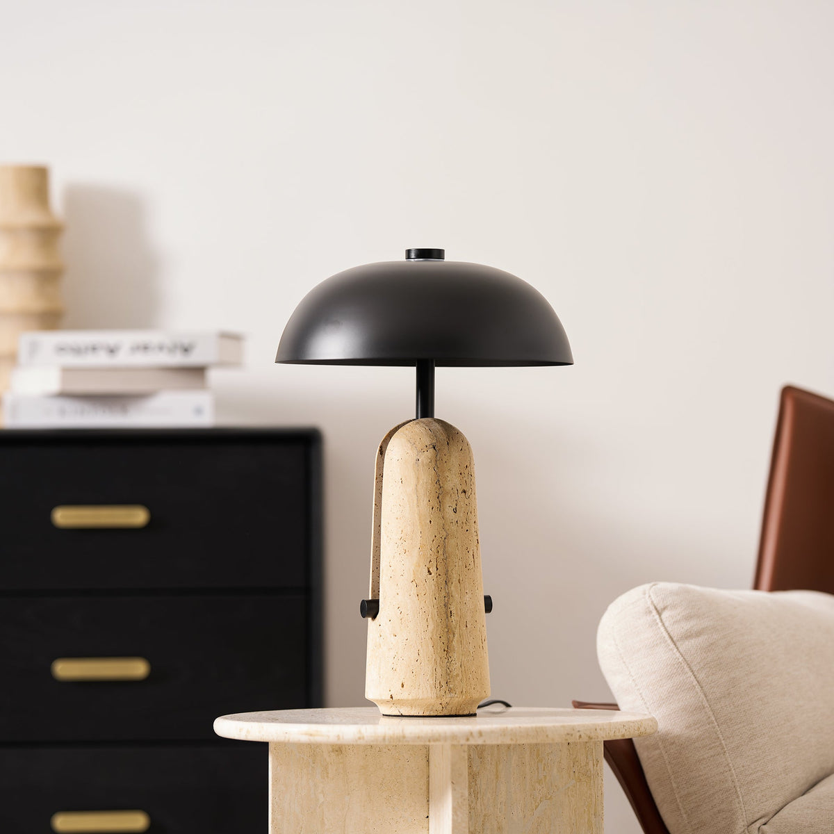 A Clérachic table lamp on a round travertine side table in front of a black chest with brass handles and a cream chair.