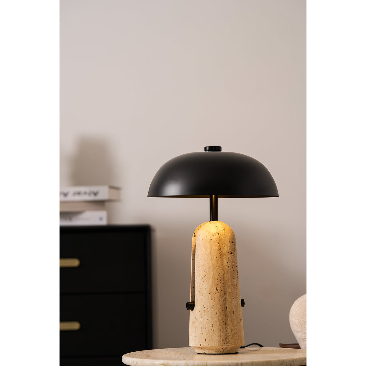 The travertine base of this Clérachic table lamp glows from within, set beside a black chest with brass handles and a book stack.