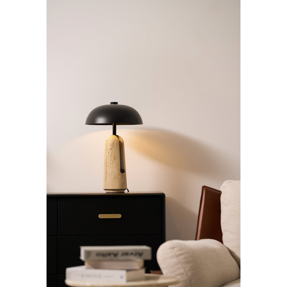 A glowing Clérachic table lamp on a black chest with brass pulls, a cream armchair and books at the lower edge.