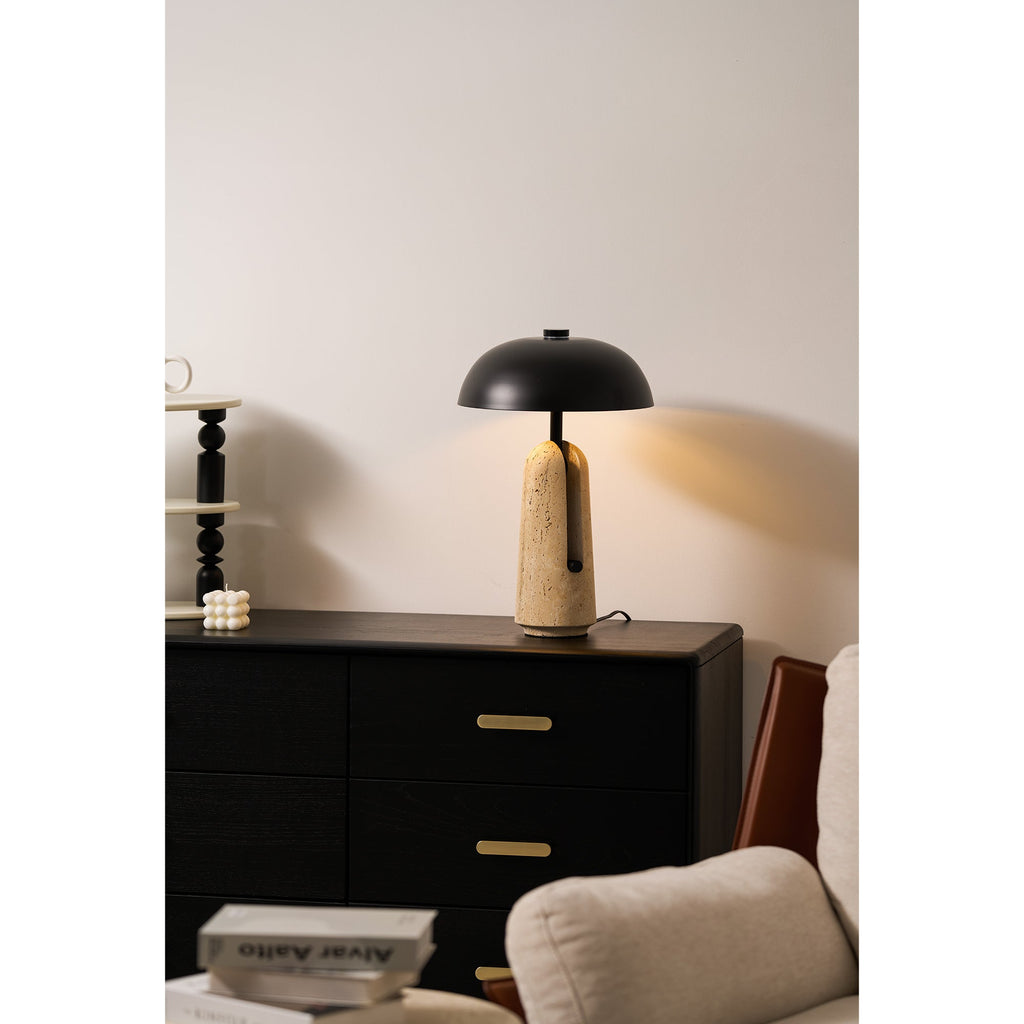 Lit Clérachic table lamp on a black chest of drawers, with a white candle, a black bead stack and a cream armchair near.