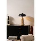 Lit Clérachic table lamp on a black chest of drawers, with a white candle, a black bead stack and a cream armchair near.