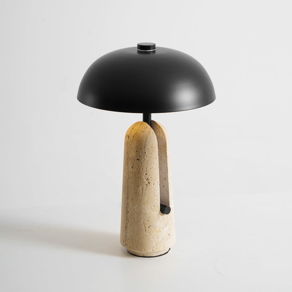 This Clérachic table lamp on a plain light grey backdrop, showing the black dome and the pitted beige travertine base.