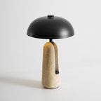 This Clérachic table lamp on a plain light grey backdrop, showing the black dome and the pitted beige travertine base.