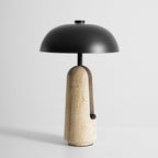 Three-quarter view of a Clérachic table lamp on a grey backdrop, with the black stem rising from a beige travertine base.