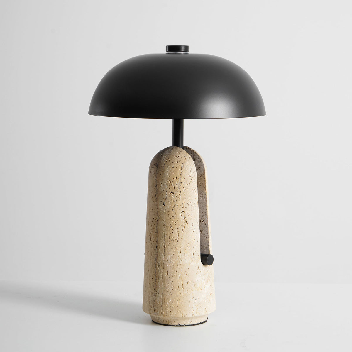 Three-quarter view of a Clérachic table lamp on a grey backdrop, with the black stem rising from a beige travertine base.