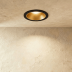Another angle of the Clérachic black and gold recessed downlight, its baffle glowing warm white overhead.