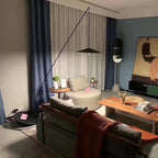 In a living room with blue curtains and a rust throw, a Clérachic floor lamp arcs from a low base to a disc shade over the seating.