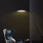 Disc shade lit warm in a dark green-black room, with a blue wing chair below the arching black stem, a Clérachic floor lamp.