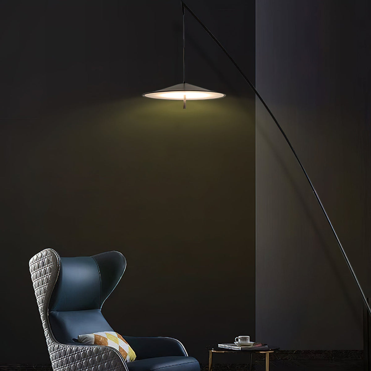 Disc shade lit warm in a dark green-black room, with a blue wing chair below the arching black stem, a Clérachic floor lamp.