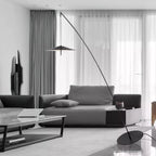 Black and white room view of a Clérachic floor lamp, its round base by a low glass coffee table and dark sofa.