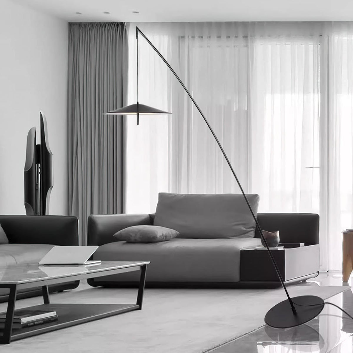Black and white room view of a Clérachic floor lamp, its round base by a low glass coffee table and dark sofa.