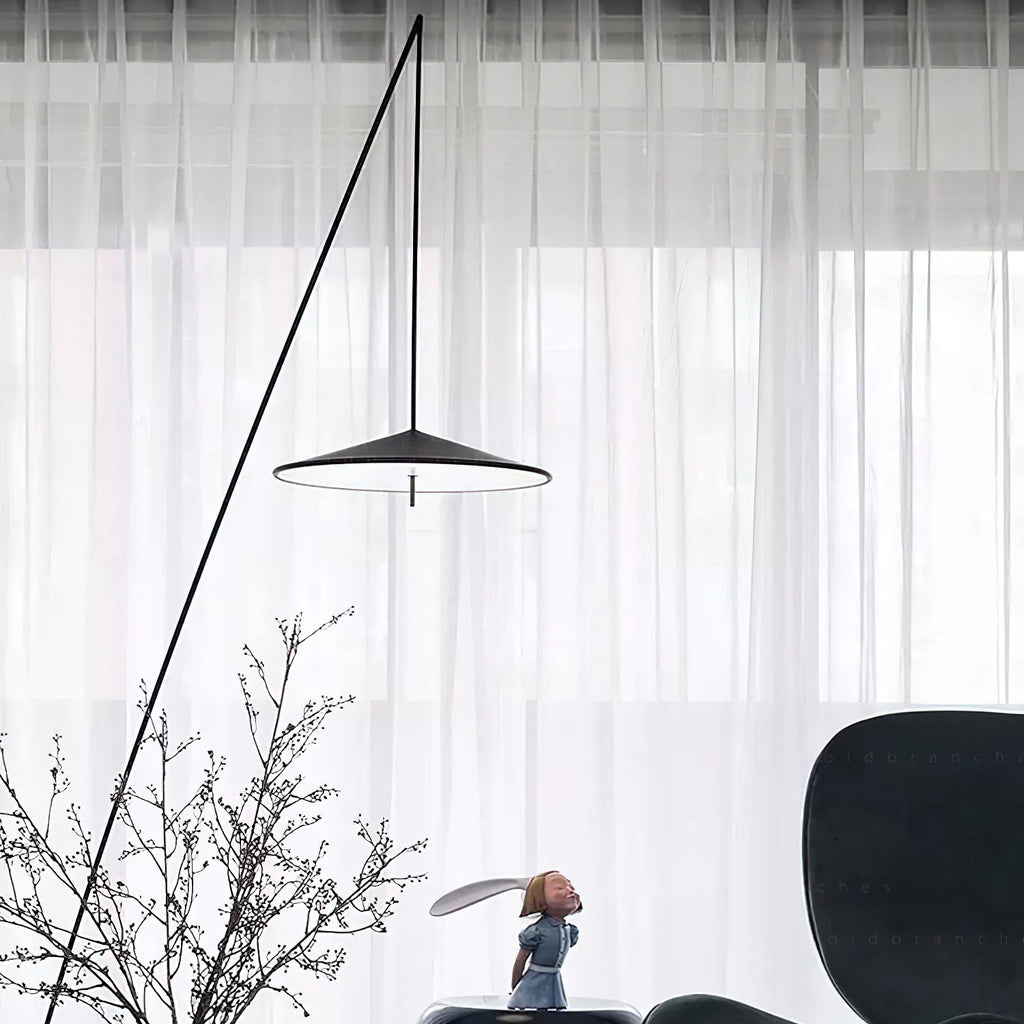 Close view of the disc shade and long diagonal stem against sheer white curtains, with bare branches in front, a Clérachic floor lamp.