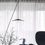 Close view of the disc shade and long diagonal stem against sheer white curtains, with bare branches in front, a Clérachic floor lamp.