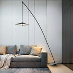 Slim black stem arcing over a grey sofa with orange and grey cushions, its round base on a wooden floor, from Clérachic.