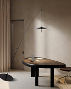 Floor lamp with a flat black base on cream flooring, its stem leaning over a dark oval table by a long curtain, from Clérachic.