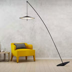 Lit disc shade hangs over a mustard armchair against a rough pale wall, the black stem curving to a tilted base, a Clérachic floor lamp.