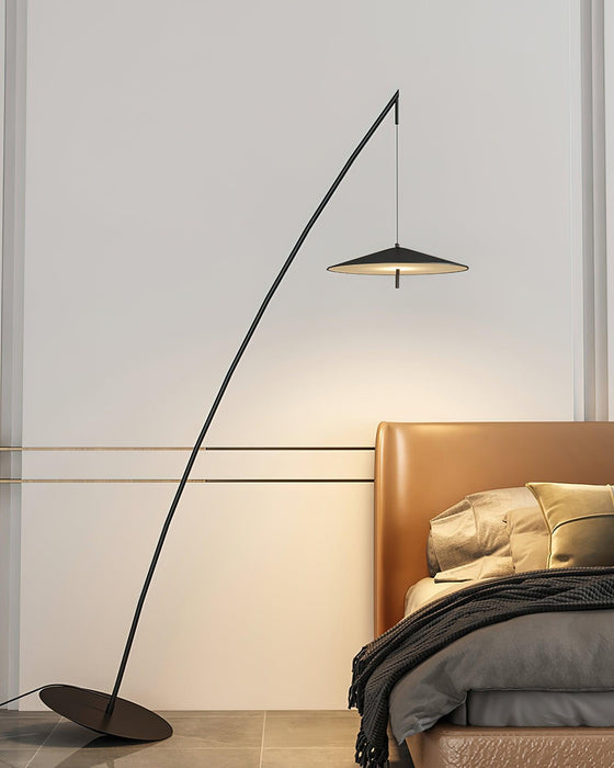 Curved black stem beside a tan leather bed, the disc shade glowing warmly against a pale wall, a Clérachic floor lamp.