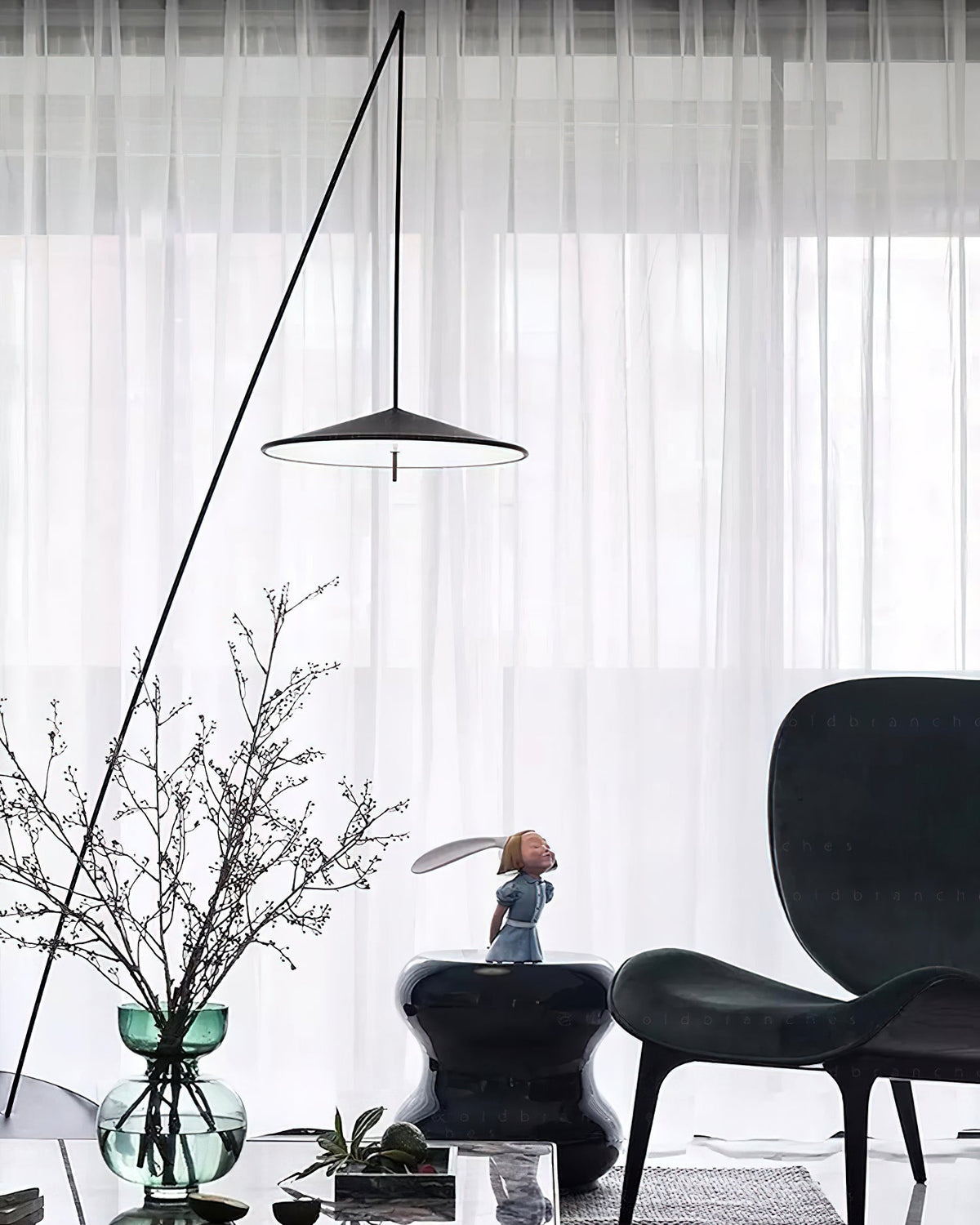 Disc shade hangs in front of white sheer curtains, with a glass vase of branches and a dark armchair below, from Clérachic.