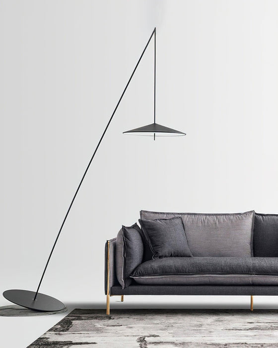 Black floor lamp on a pale floor beside a dark grey sofa with gold legs, its round base angled slightly, a Clérachic floor lamp.