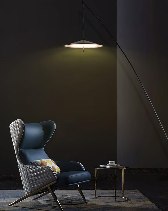 Tall portrait view of a lit disc shade above a blue wing chair in a dark room, with the stem curving to the right, from Clérachic.
