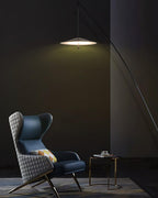 Tall portrait view of a lit disc shade above a blue wing chair in a dark room, with the stem curving to the right, from Clérachic.