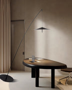 Tall view of the black stem sweeping over a dark oval desk, with beige curtains and a tilted flat base, a Clérachic floor lamp.