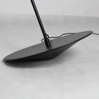 Close view of the round black base, tilted on a concrete-grey floor, with the stem and cord leaving it, on this Clérachic floor lamp.