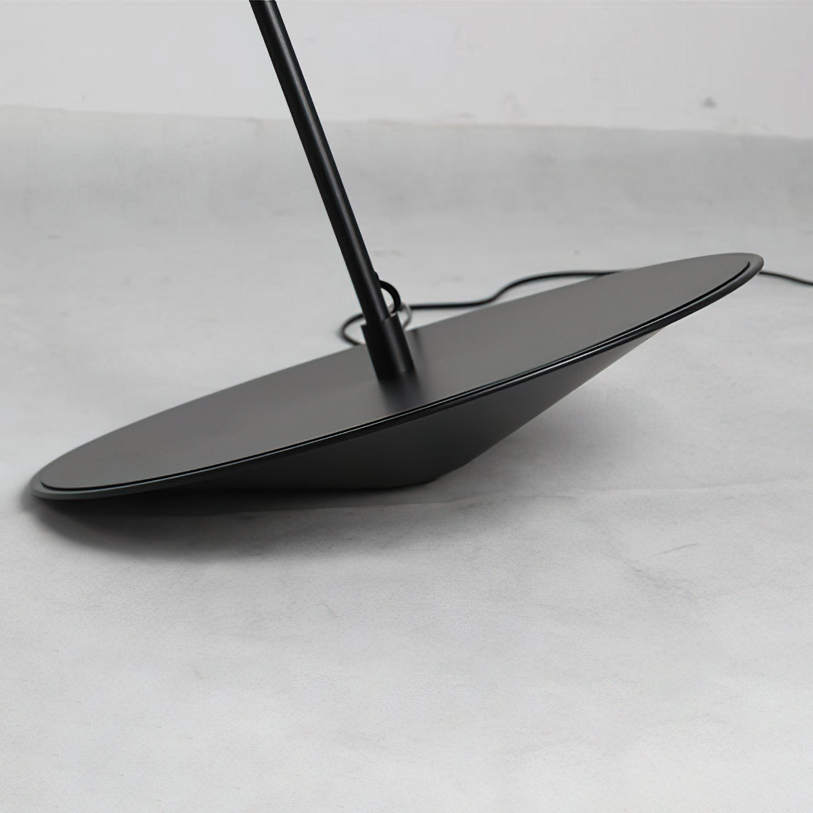 Close view of the round black base, tilted on a concrete-grey floor, with the stem and cord leaving it, on this Clérachic floor lamp.