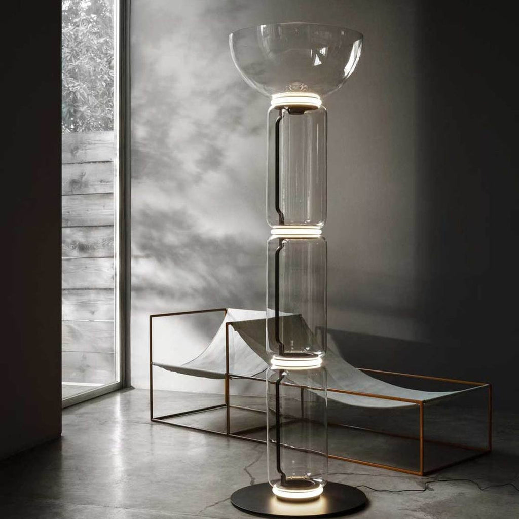 Tall Clérachic floor lamp with a glass bowl on top, glowing in a concrete-floored room beside a low copper-framed lounger and a tall window.