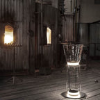 Clérachic floor lamp with a flared glass top standing in a dim, rusty industrial space, its base ring glowing on a worn wooden floor.