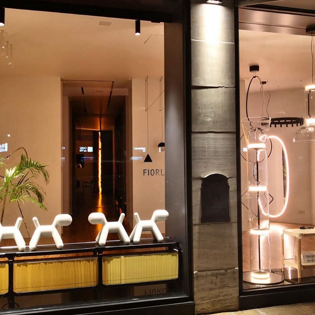 Shop-window view at night with a Clérachic floor lamp among other lights, beside white dog-shaped stools and a leafy plant inside.