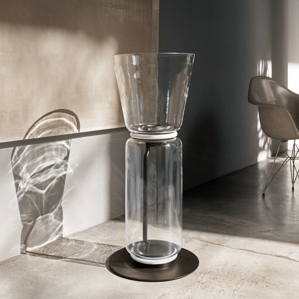 A Clérachic floor lamp with a flared glass top on a round black base, in a pale concrete room with sunlight casting a shadow on the wall.