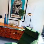 Part of a Clérachic floor lamp with a tall flared glass top, beside a burr-wood sideboard with blue bottles, a small tree and a framed poster.