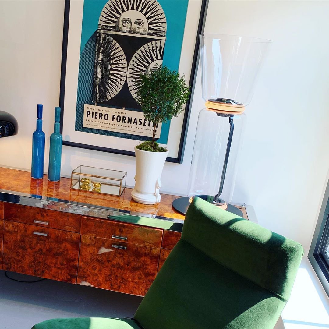 Part of a Clérachic floor lamp with a tall flared glass top, beside a burr-wood sideboard with blue bottles, a small tree and a framed poster.