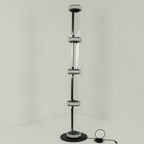 Unlit Clérachic floor lamp of four glass cylinders strung on a thin black stem, with a small control on the floor, in a pale corner.