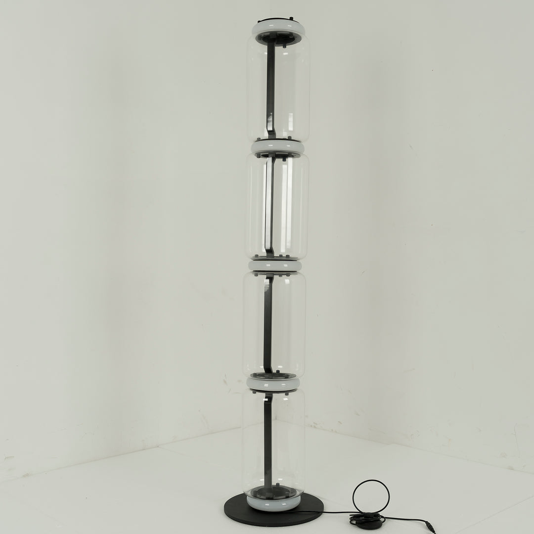 Unlit Clérachic floor lamp of four glass cylinders strung on a thin black stem, with a small control on the floor, in a pale corner.