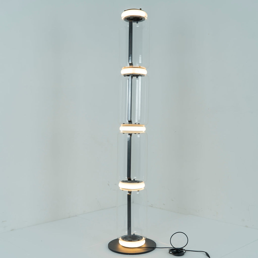 Clérachic floor lamp lit in a pale corner, a ring of white light glowing at each join between its four glass cylinders.