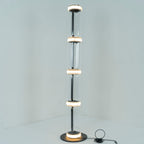 Clérachic floor lamp lit in a pale corner, a ring of white light glowing at each join between its four glass cylinders.