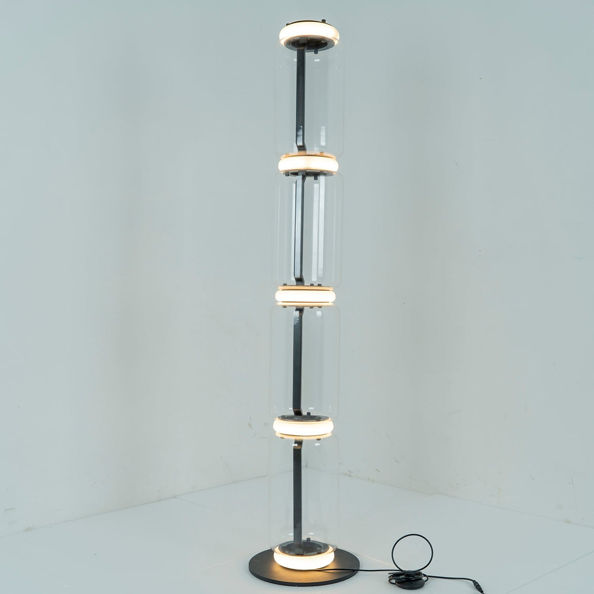 Clérachic floor lamp lit in a pale corner, a ring of white light glowing at each join between its four glass cylinders.
