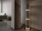 A Clérachic floor lamp with a flared glass top, lit on a concrete floor beside a pale column, with a sofa in the soft light behind.