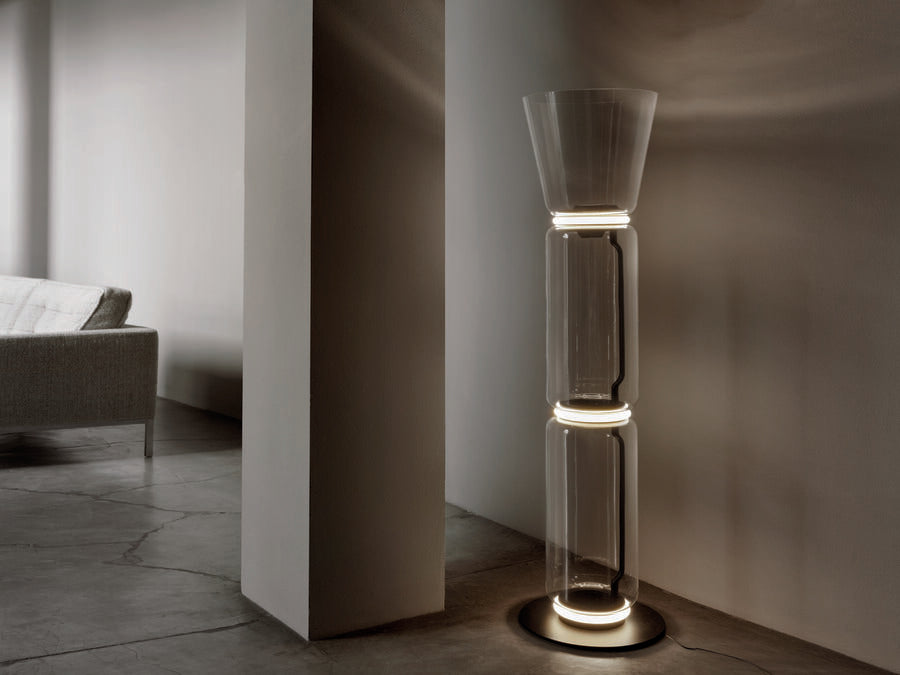A Clérachic floor lamp with a flared glass top, lit on a concrete floor beside a pale column, with a sofa in the soft light behind.