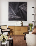 Clérachic floor lamp at the right edge of a living room with a dark abstract painting, a brass sideboard and a cane-backed chair.