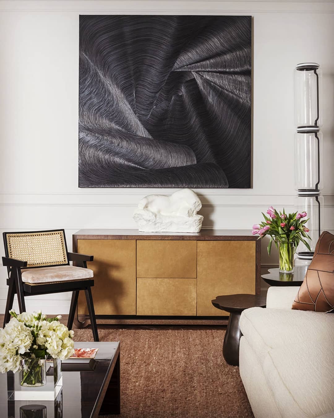 Clérachic floor lamp at the right edge of a living room with a dark abstract painting, a brass sideboard and a cane-backed chair.
