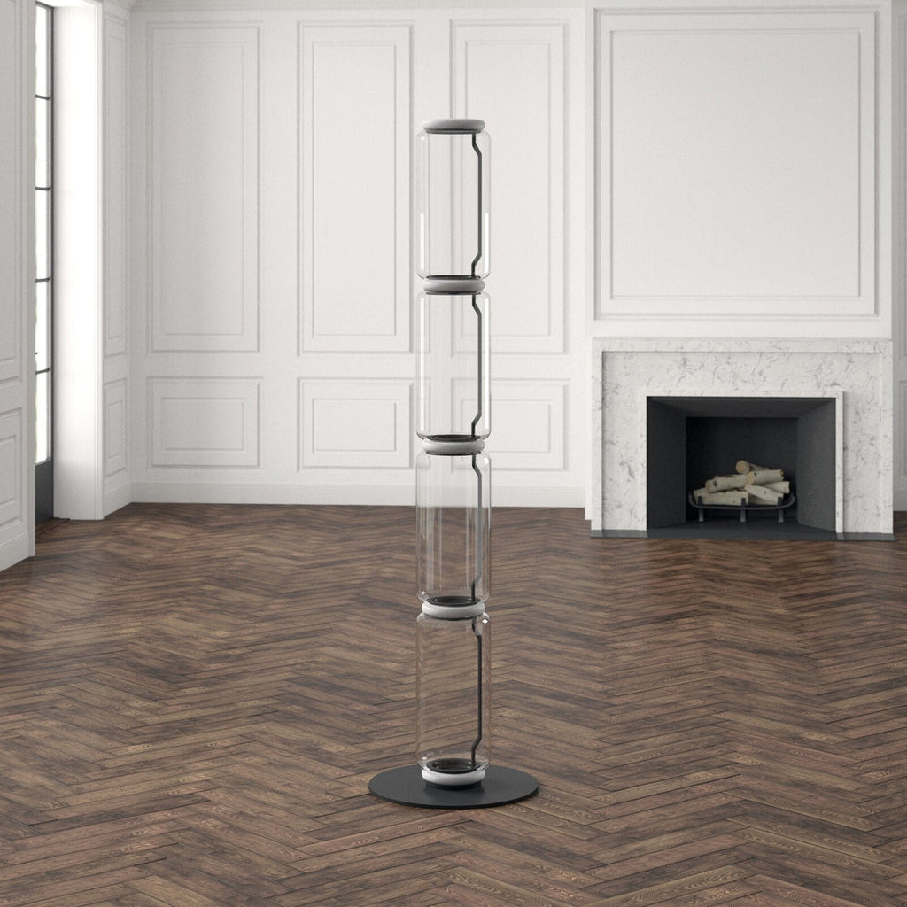 Unlit Clérachic floor lamp of four stacked glass cylinders in a white panelled room with a marble fireplace and herringbone floor.