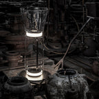 Clérachic floor lamp with a flared glass top, lit among old iron machinery in a dark industrial room.
