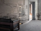 Clérachic floor lamp lit in a worn plaster room with a tall doorway, with a black leather armchair in front.