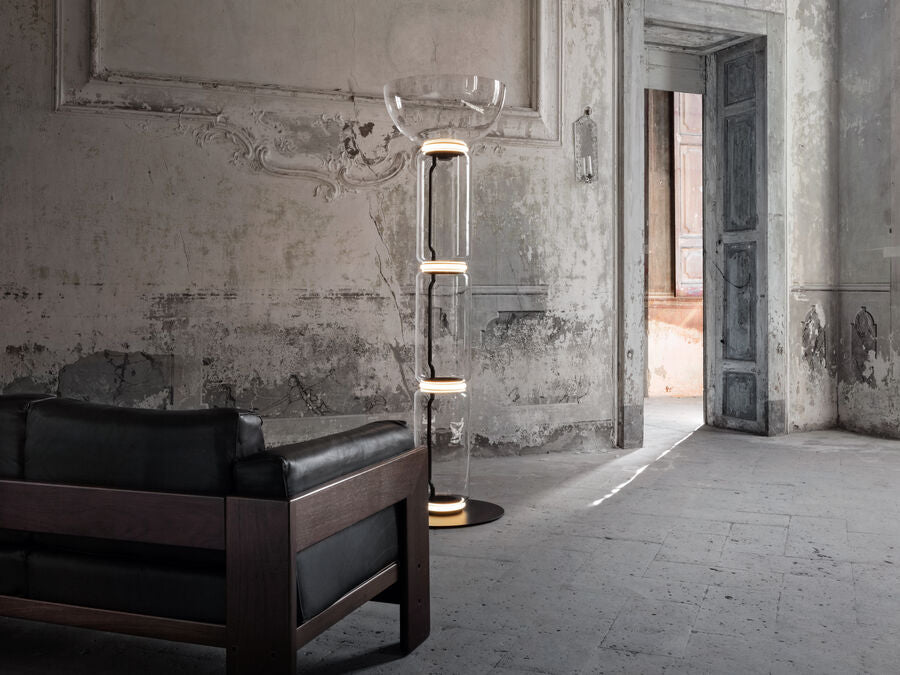 Clérachic floor lamp lit in a worn plaster room with a tall doorway, with a black leather armchair in front.