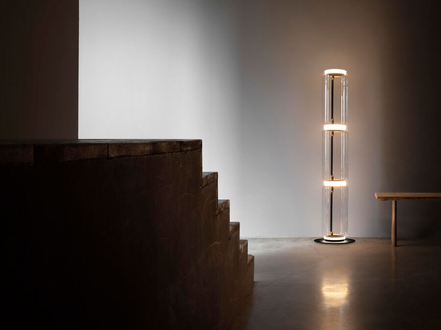 Distant Clérachic floor lamp glowing in a bare, light-toned room beside a dark wooden staircase and a low bench.